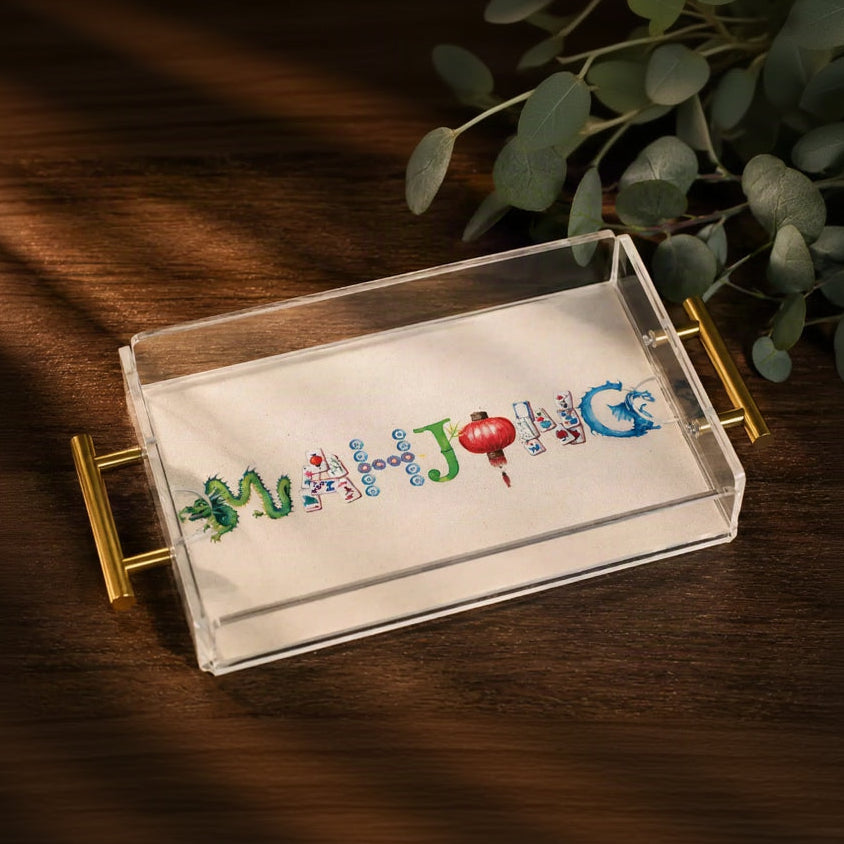 A clear acrylic rectangular tray with gold metal handles, containing a white sheet printed with colorful Chinese characters spelling 'Merry Joy' in green, blue, red, and gold tones. The tray is placed on a dark wooden table with eucalyptus leaves in the background.