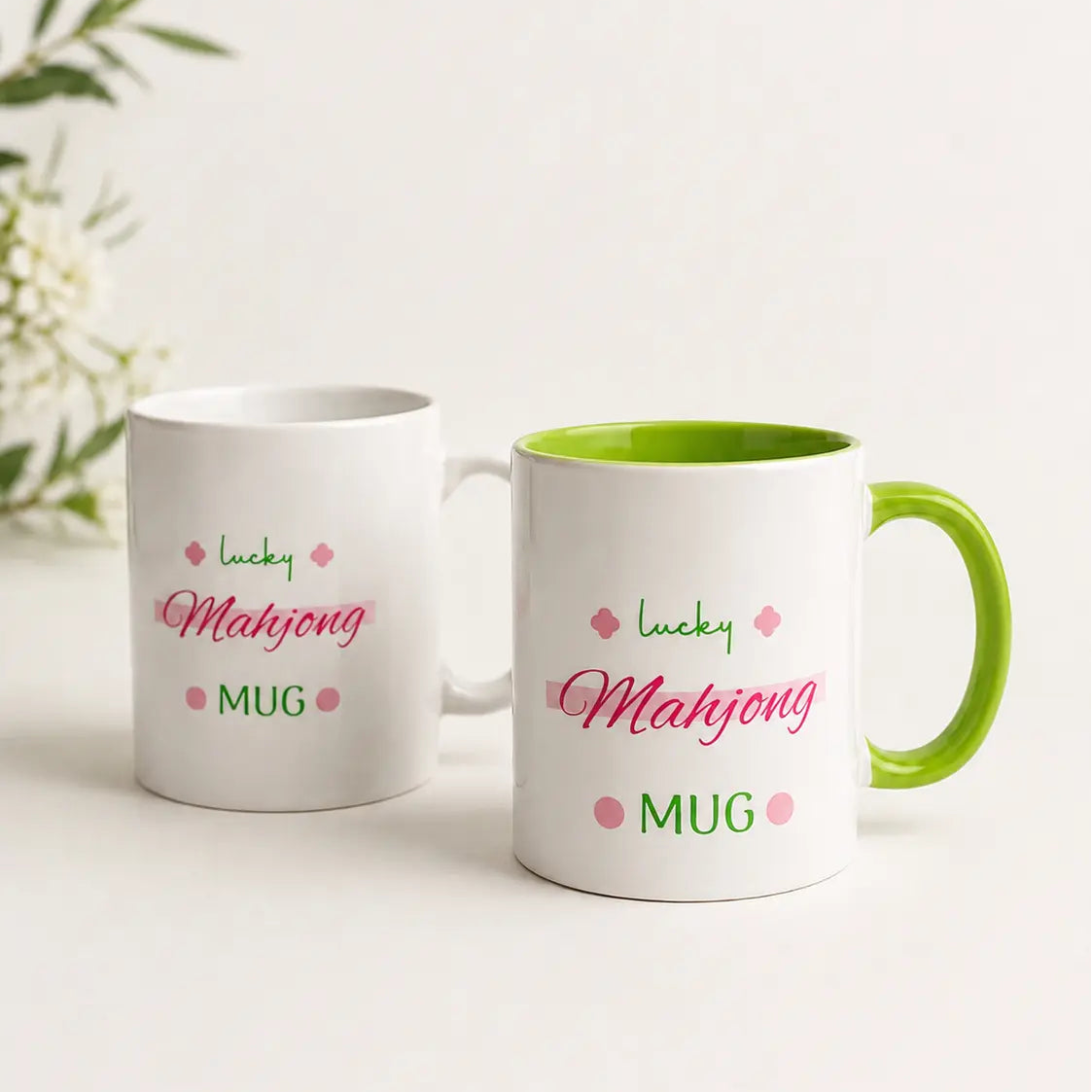 Two white Organiks Plus ceramic mugs with green handles and interiors, printed with pink and green text reading 'lucky Mahjong MUG' on a clean white background.
