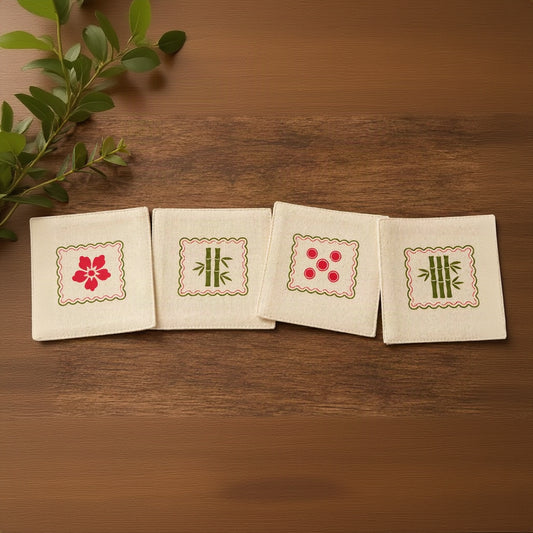 Four square cream fabric coasters with embroidered Japanese motifs, arranged in a row on a dark wood tabletop with a small green leaf sprig in the top-left corner.
