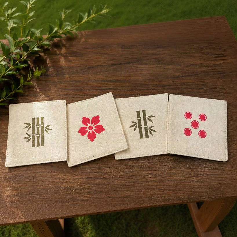 Four square cream fabric coasters with embroidered Japanese-style designs, including bamboo, a red flower, and red dots, arranged on a dark wood surface.