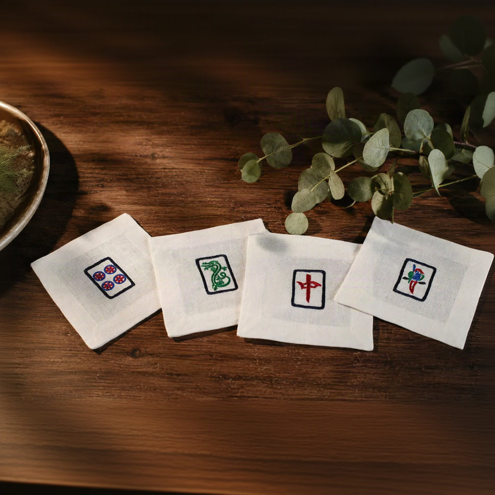 Four white square cotton napkins with embroidered Chinese mahjong tile designs arranged on a dark wooden table next to green eucalyptus leaves.
