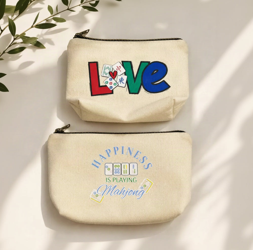 Two cream Organiks Plus canvas zip pouches displayed on a light beige background with soft leaf shadows. The top pouch features the word 'LOVE' in colorful block letters with playing card details, and the bottom pouch reads 'HAPPINESS IS PLAYING Mahjong' with matching playing card graphics.