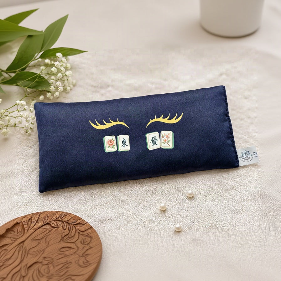 A navy blue eye pillow featuring embroidered Chinese mahjong tile graphics and gold eyelashes, styled on a textured cream fabric.