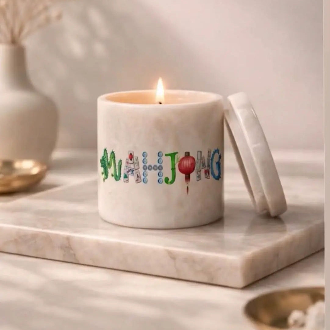 A white Organiks Plus marble jar candle with a lit flame sits on a matching marble tray, with its lid leaning against the right side. The front of the candle is decorated with colorful lettering spelling 'MAHJONG' in green, blue, and red, with small festive accents.