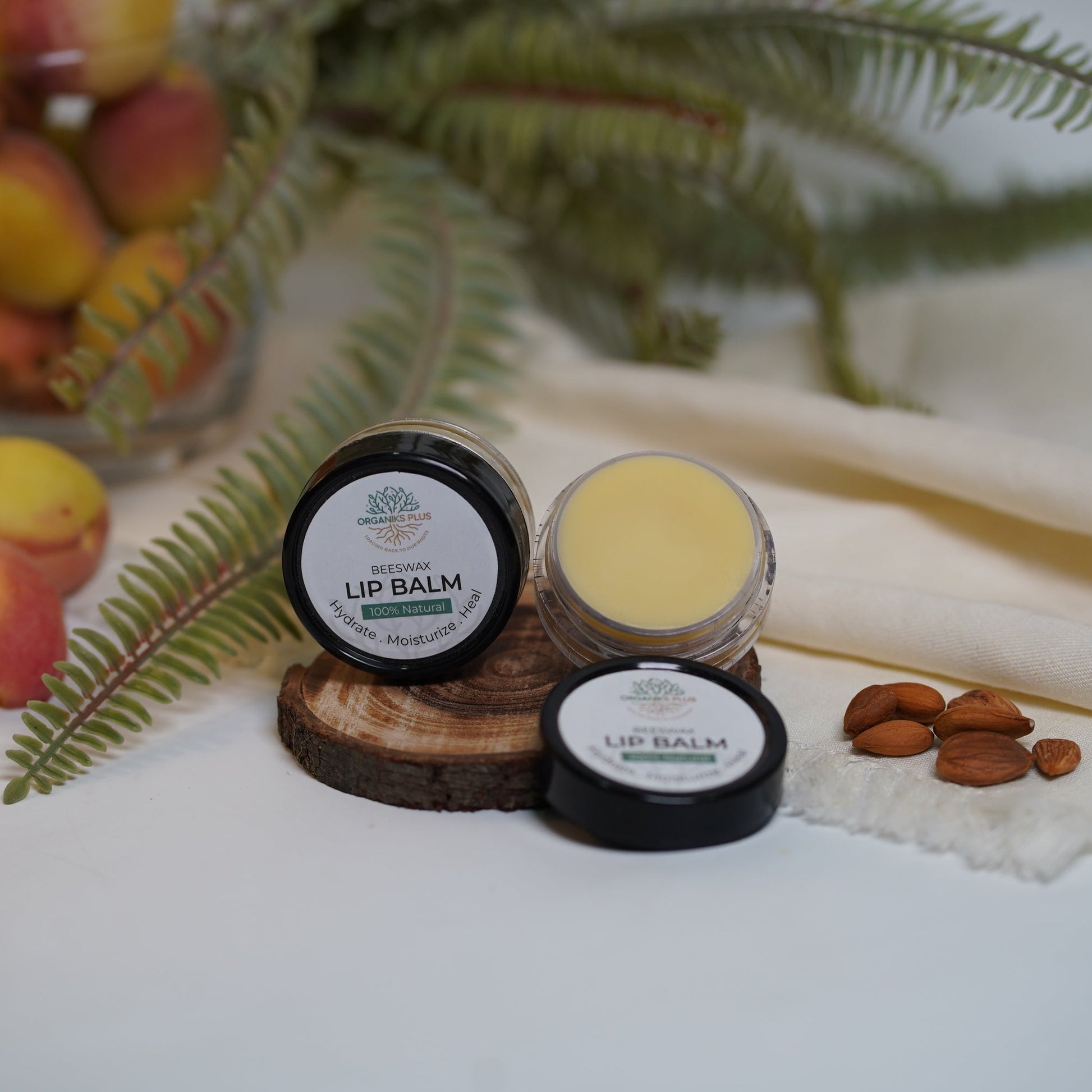 lip balm containers on a white surface with fruits and a plant in the background


