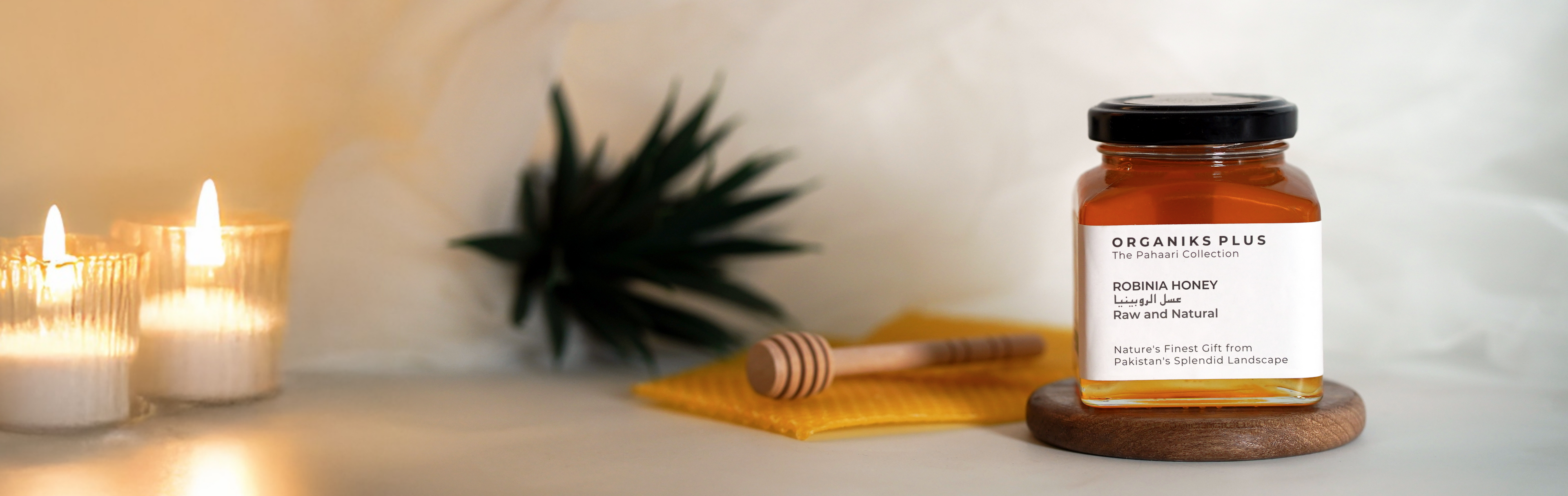 Jar of honey with a label on a wooden surface next to lit candles and a plant.

