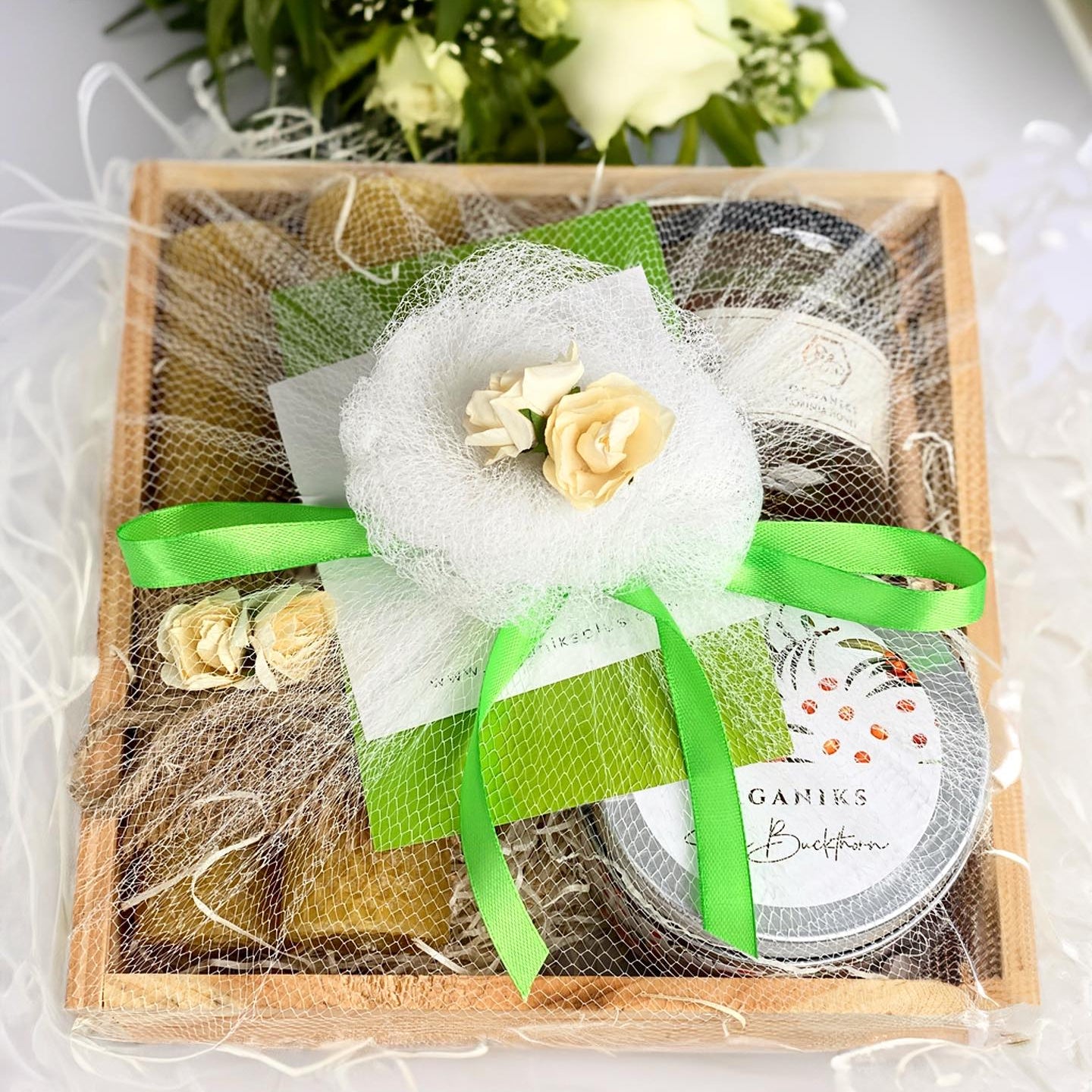 A wellness gift pack containing a wooden tray, a jar of herbal tea, a beeswax candle, and a small bouquet of flowers, all tied with green ribbons and white fabric.