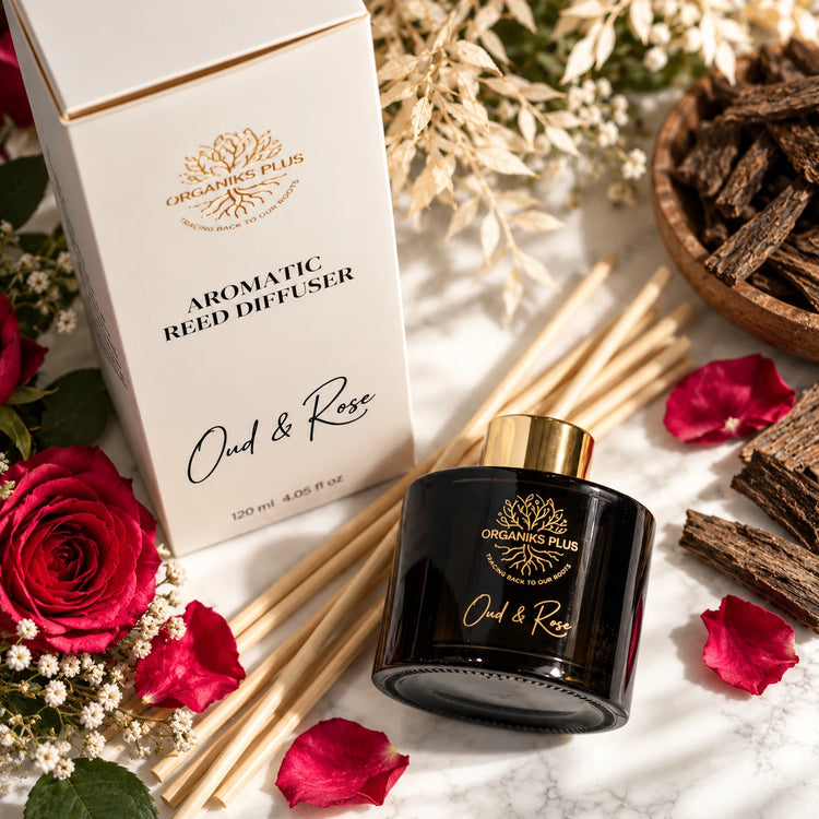 Organiks Plus Oud and Rose aromatic reed diffuser in a dark glass bottle with gold foil logo, cream packaging with black lettering, natural reeds, red roses and oud wood on a marble surface